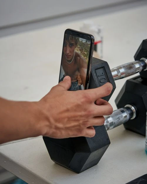 A smartphone shows a person's portrait as it balances on a black dumbbell with a hand touching it, other dumbbells nearby.