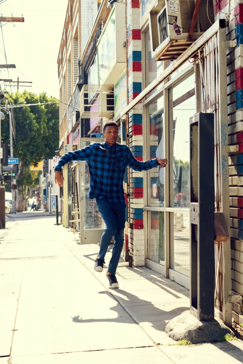 Man in blue flannel shirt leaps on city sidewalk past colorful storefronts on sunny day, captured in medium shot. Urban street scene.