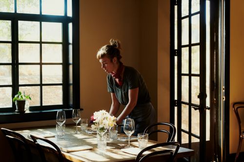 A woman arranges a dining table with wine glasses, flowers, and napkins next to a window, bathed in soft, natural light in an indoor setting.