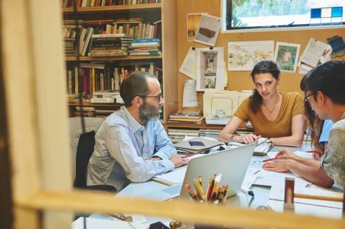 Four people, including an adult woman and man, collaborate around a table with a laptop in a bright indoor space filled with books.