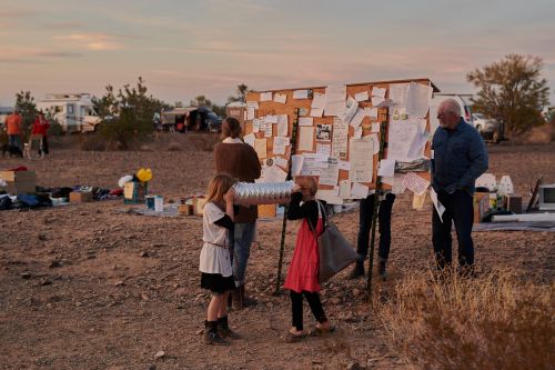 A group of people, including two children, stand around a bulletin board covered with papers in a dry, open landscape at sunset.