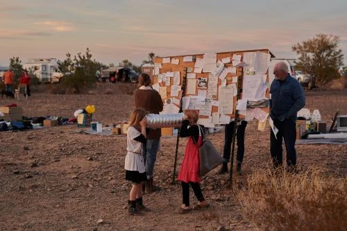 A group of people, including two children, stand around a bulletin board covered with papers in a dry, open landscape at sunset.
