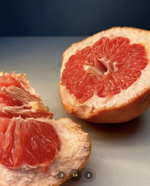 Halved grapefruit with visible red pulp and a thick rind sits on a white surface against a dark blue-gray background. Close-up food shot.