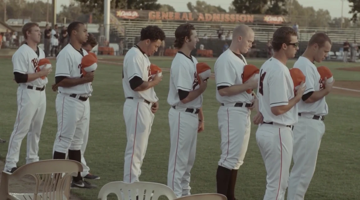 Baseball team stands on field in uniforms with baseball hats, facing forward, during daytime with stadium seating in the background.