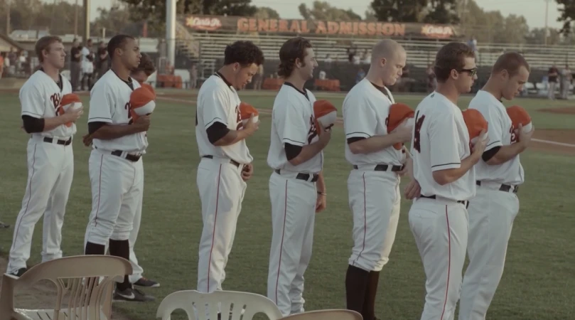 Baseball team stands on field in uniforms with baseball hats, facing forward, during daytime with stadium seating in the background.