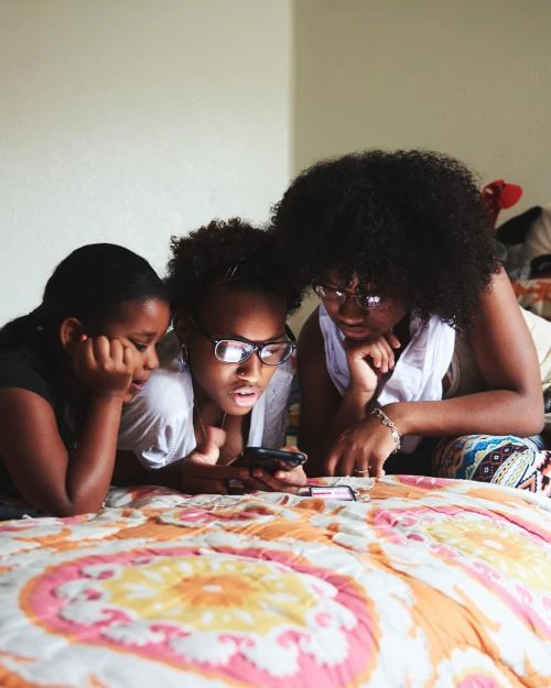 Three dark-haired teenage girls lying on a bed using a smartphone, wearing casual clothing, with a cream-colored wall in the background.