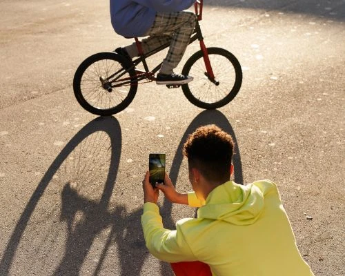 Person on bike casts shadow as another person photographs them with a phone outdoors on an asphalt surface in bright daylight.