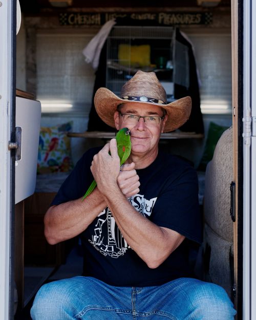 Portrait of a man in a cowboy hat holding a green parrot inside a camper van. He's wearing glasses and a black t-shirt with a graphic.
