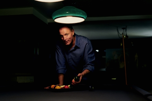 Man in blue shirt lines up billiard balls on a pool table under a green-shaded lamp in a dimly lit room, focused and ready to play.