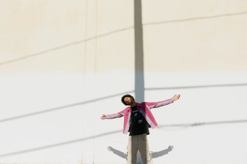 Man in a pink jacket and light trousers standing with arms outstretched against a white wall with a vertical shadow.