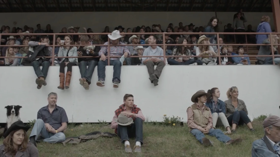 Outdoor crowd scene shows people in stadium seating and on grassy hillside. The group watches an event from an eye-level medium shot.