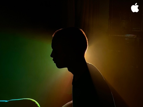 Silhouette of a young person with short hair, lit with yellow and green light in a dark indoor setting, with an Apple logo visible.