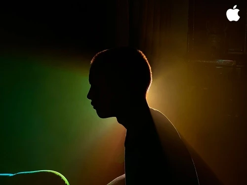 Silhouette of a young person with short hair, lit with yellow and green light in a dark indoor setting, with an Apple logo visible.