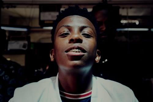 Portrait of a smiling young Black male with braces wearing a white coat in a dark indoor setting with a blurred background.