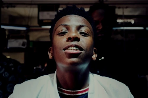 Portrait of a smiling young Black male with braces wearing a white coat in a dark indoor setting with a blurred background.