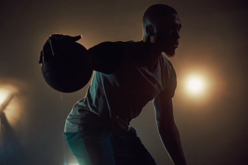 Silhouette of a man dribbling a basketball, captured with moody lighting, creating a dramatic sports action shot in a dark indoor setting.