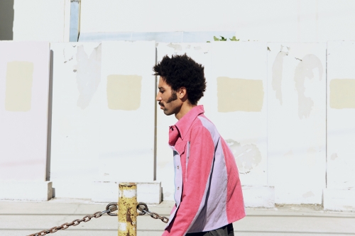 Side profile of a stylish adult male with an afro and mustache wearing a pink jacket while walking outdoors on a sunny day.