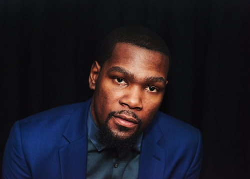 Portrait of an adult Black man wearing a blue blazer and black shirt, looking at the camera against a dark background.