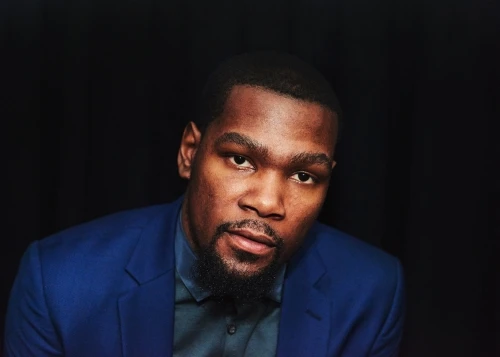Portrait of an adult Black man wearing a blue blazer and black shirt, looking at the camera against a dark background.