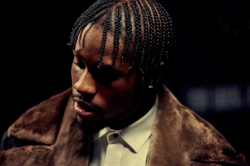 Portrait of a stylish Black man with cornrows wearing a fur coat indoors against a dark background, looking away from camera.