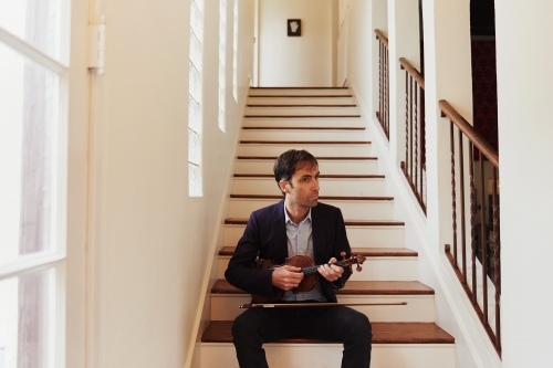 Man sits on stairs playing a violin in a brightly lit indoor setting, wearing a dark blazer and black pants, with a focused expression.