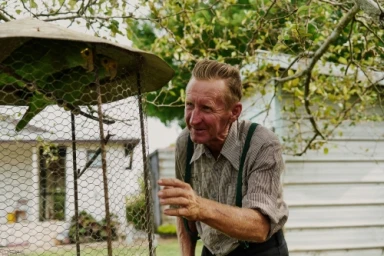 Elderly man with suspenders interacting with a green parrot inside an outdoor wire birdcage by photographer Jake Michaels