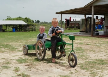 Two young boys sitting on a green homemade pedal cart in a grassy farm yard with a building in the background.