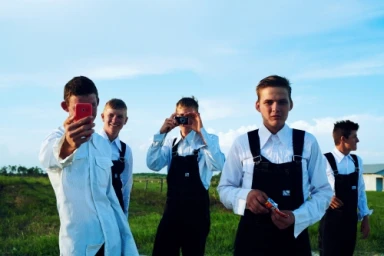 Five young men in white shirts and black overalls standing in a grassy field under a bright blue sky.
