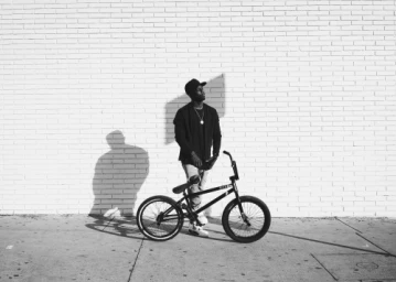 A Black man wearing a cap standing beside a BMX bicycle against a white painted brick wall.