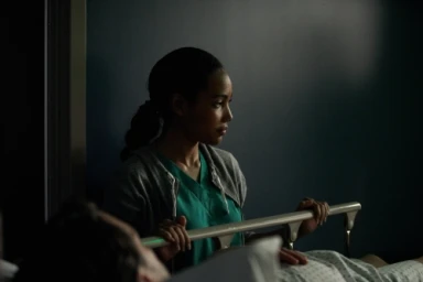 A Black woman in medical scrubs and grey hoodie standing by a hospital bed in low key lighting, photographed by Jake Michaels.