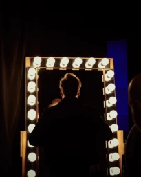 Silhouetted person facing an illuminated vanity mirror framed with glowing round light bulbs in a dark room