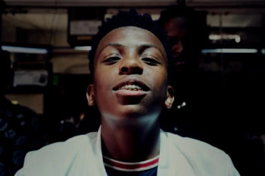 Close-up portrait of a young Black man looking down at the camera with gold grills on his teeth, wearing a white collared shirt.