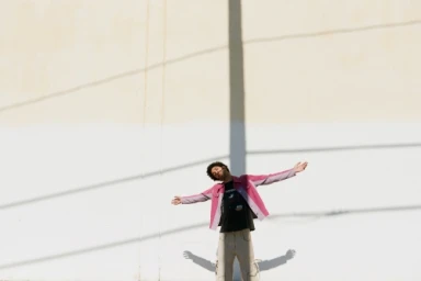 Man in a pink jacket and light trousers standing with arms outstretched against a white wall with a vertical shadow.