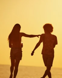 Silhouette of a man and woman walking on a beach at sunset, holding a glass bottle against a warm glowing sky.