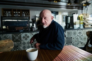 Elderly man with white facial hair wearing a dark jacket seated at a wooden slatted table inside a café.