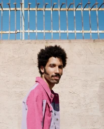 Man with afro and mustache wearing a pink corduroy jacket against a stucco wall and blue sky