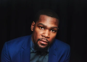 Portrait of Kevin Durant wearing a blue suit jacket and dark shirt against a dark background, photographed by Jake Michaels.