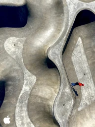 High-angle aerial view of a skater in a red top inside a sweeping concrete skatepark bowl with curves.