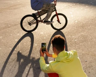 Man in neon yellow hoodie crouches on asphalt to photograph a person on a bicycle against the bright sunlight.