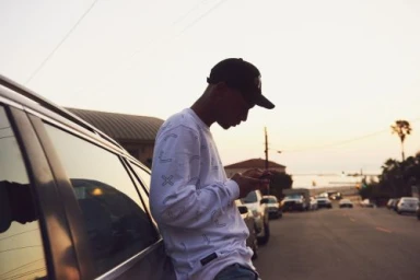 Young man wearing a patterned long-sleeve white shirt and black baseball cap leaning against a car on a street at sunset.