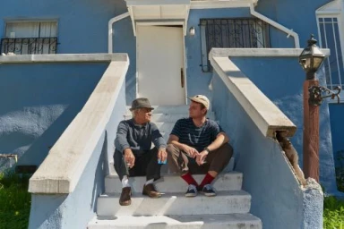 Two men sitting and talking on concrete steps outside a blue residential building in Los Angeles.