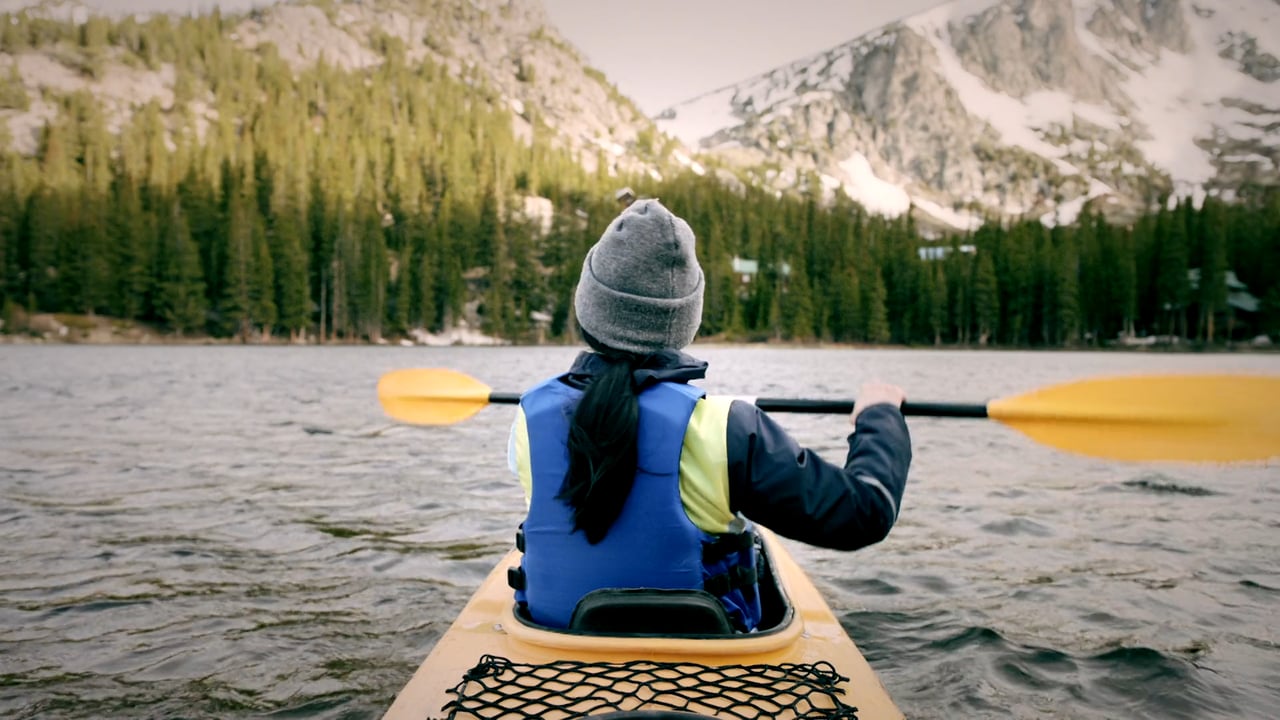 A person kayaking on a lake surrounded by mountains and evergreen trees. The kayaker is seen from the back and wearing a hat.