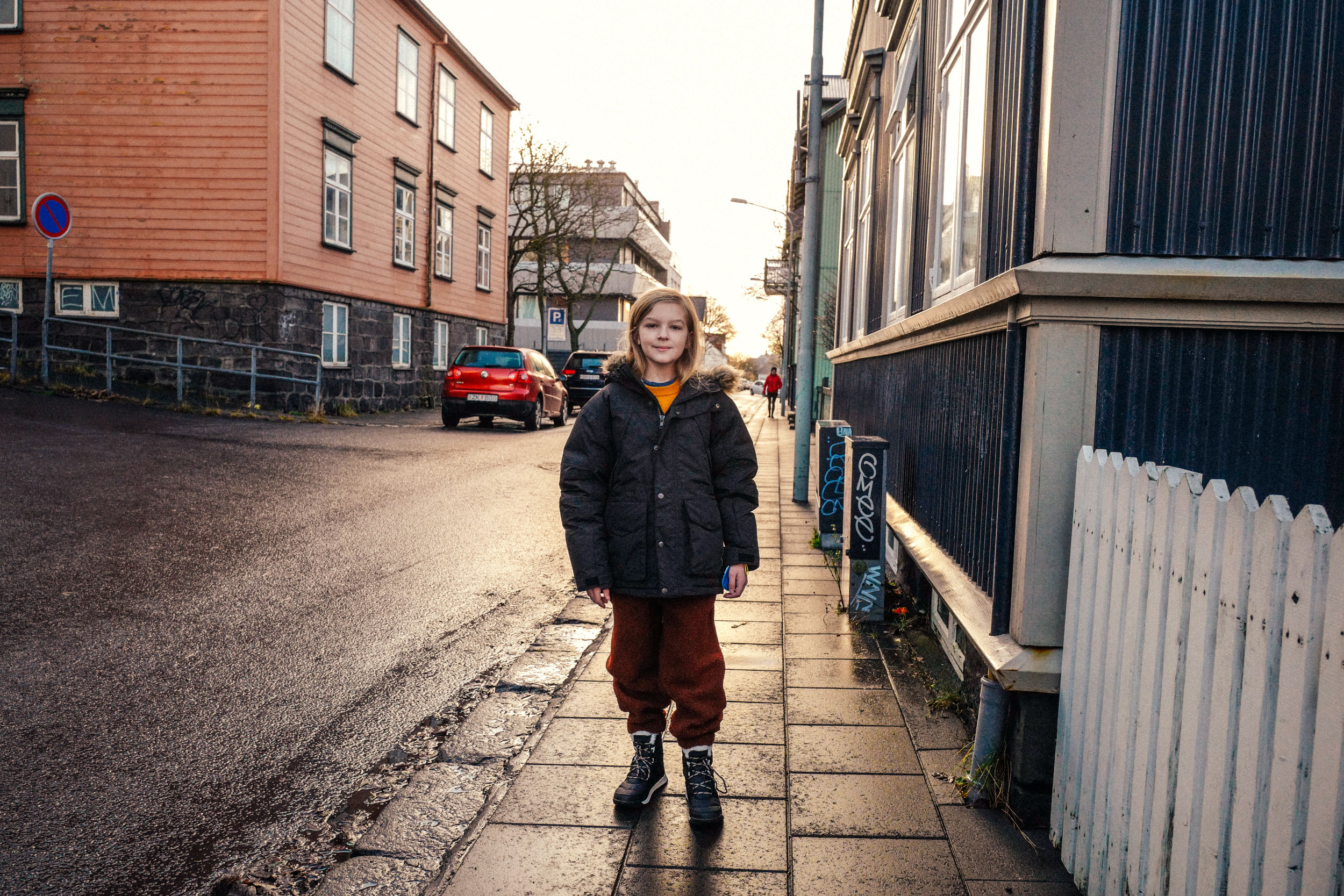A young girl in a winter coat poses on a sidewalk in a European city, with colorful buildings and parked cars in the background.