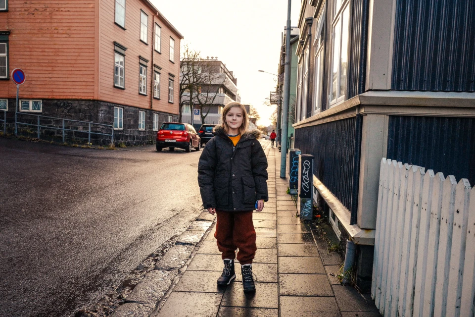 A young girl in a winter coat poses on a sidewalk in a European city, with colorful buildings and parked cars in the background.