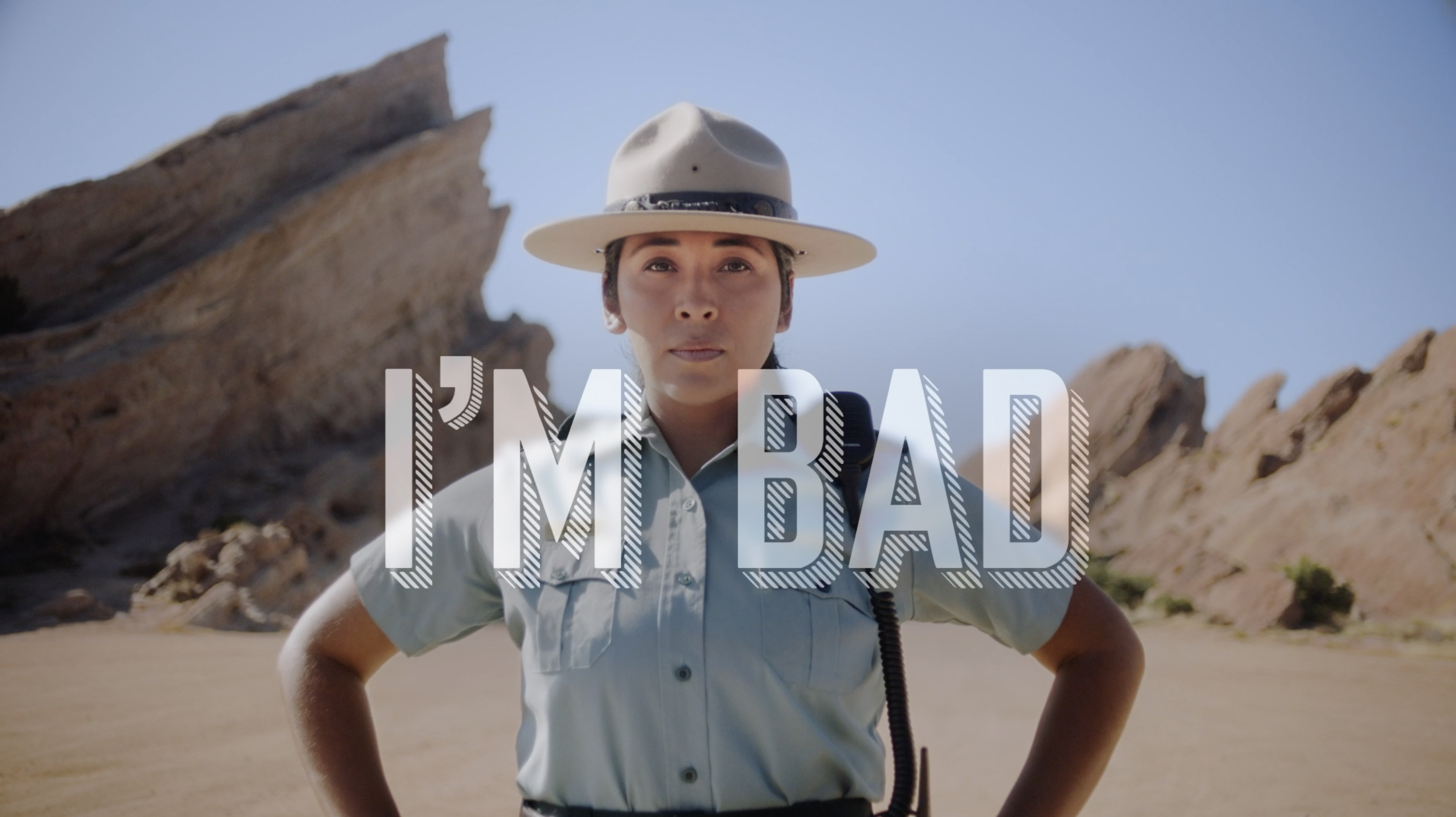 Female park ranger in hat poses in a desert landscape with rock formations under a clear sky, overlaid with the text 'I'M BAD'.