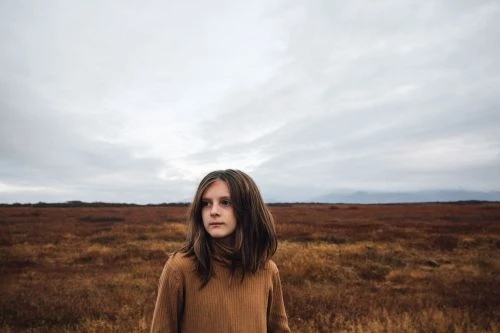 Portrait of a young person in a brown sweater standing in a field of brown grass under a cloudy sky. Landscape orientation.