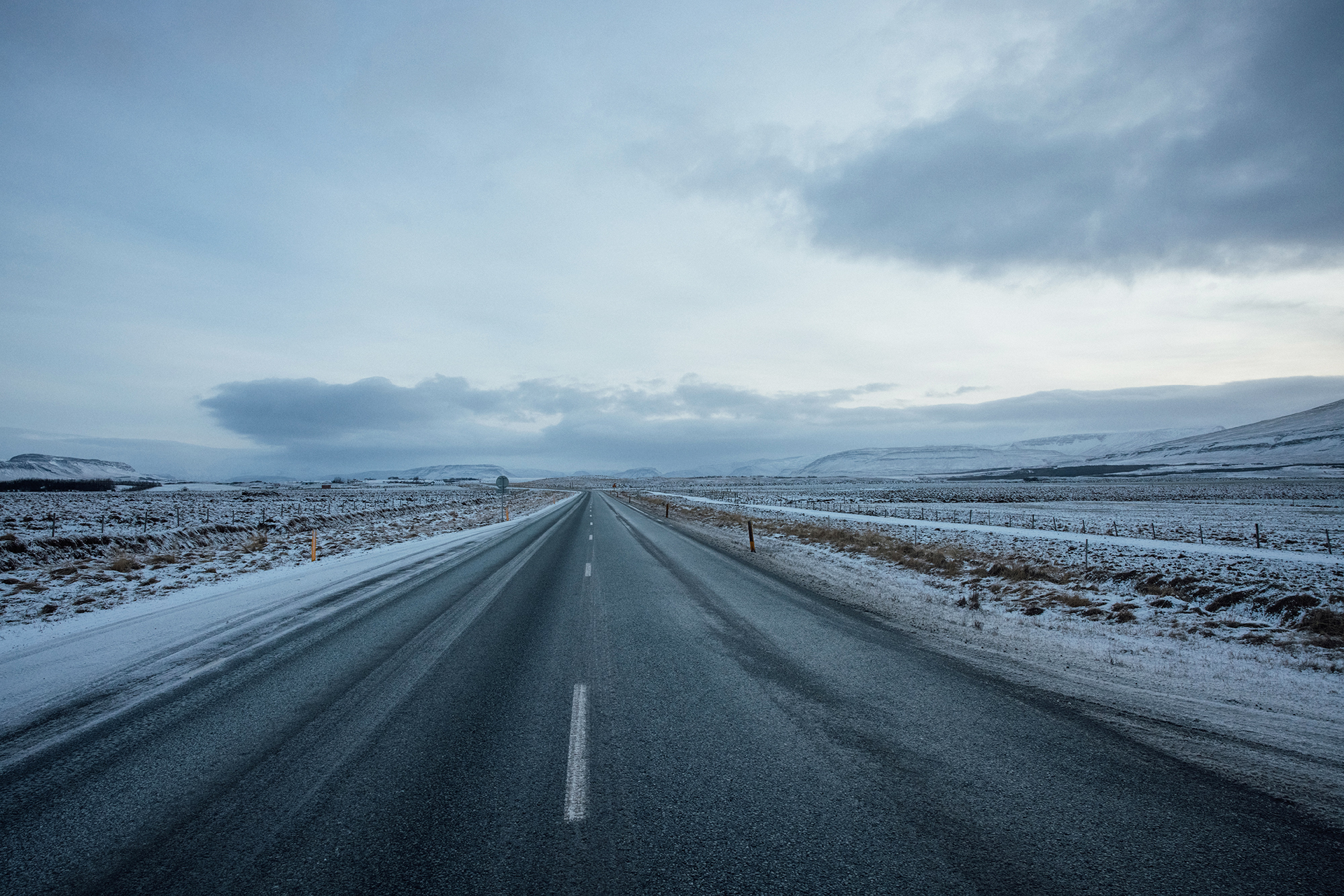 A long asphalt road stretches through a snowy landscape under a cloudy sky, creating a serene and isolated travel scene.