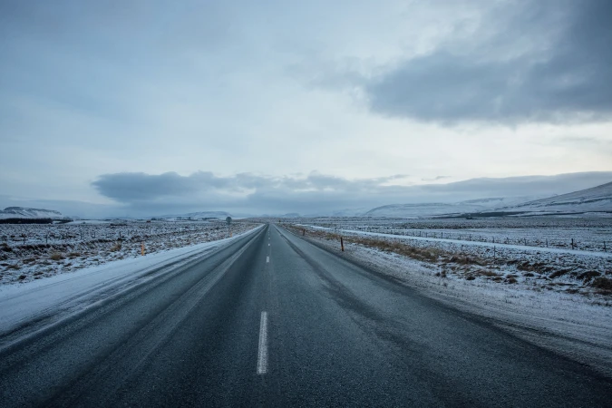 A long asphalt road stretches through a snowy landscape under a cloudy sky, creating a serene and isolated travel scene.