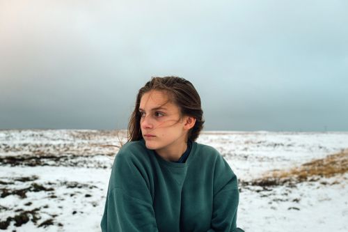 Portrait of a young woman with long hair looking left, wearing a teal sweater, against a snowy landscape and overcast sky.
