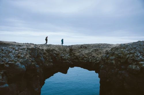 Two people walk on a rocky cliff formation with a natural arch over the ocean under a cloudy sky, captured in a wide shot.